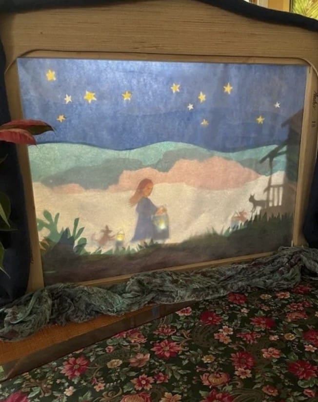Image of a colorful shadow puppet stage showing a girl with a lantern walking towards a cabin. Stars fill the sky.